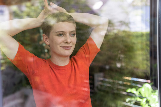 Close-up of thoughtful woman with arms raised standing at home seen through window