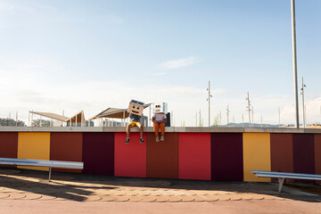 Friends wearing robot costumes sitting on colorful retaining wall against sky during sunny day
