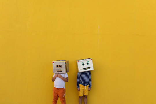 Boys wearing robot masks made of boxes while standing against yellow wall