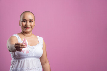 Mujer sonriente con cáncer de mama en fondo rosado mostrando un lazo rosa - obrazy, fototapety, plakaty