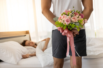 Surprised husband hiding bouquet of flower behind his back before surprise his wife. Beautiful romantic couple in bed room