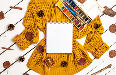 Flat lay of sketchbook, paints with brushes, yellow sweater, leaves, cinnamon sticks and pine cones on a white wooden background. Autumn composition with place for text.