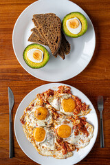 Close up fresh avocado cut in half, slices of bread and fried eggs with tomato on white plates on wooden table