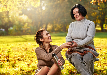 Portrait of beautiful young woman drinking tea with her mom in autumn park