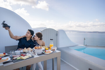 Couple having breakfast, tourist take a selfie on terrace hotel outdoor. Luxury and delicious food. Santorini, Greece.
