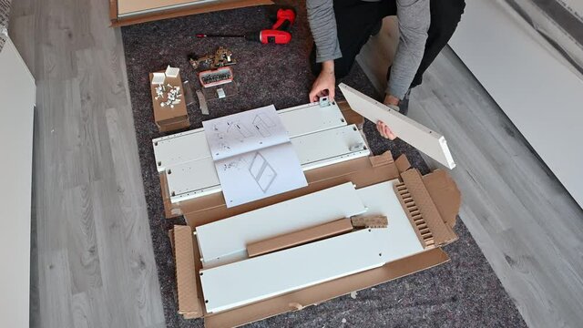 Scenes in the house: assembling a piece of furniture, the Caucasian man interlocks two boards that make up the piece of furniture. Next to the cabinet assembly manual.