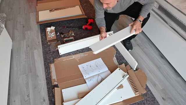 Scenes in the house: assembling a piece of furniture, the Caucasian man interlocks two boards that make up the piece of furniture. Next to the cabinet assembly manual.