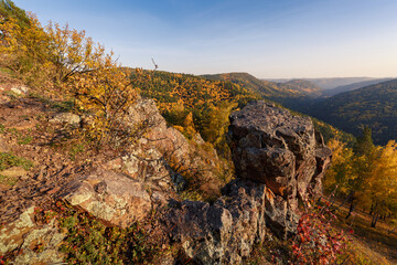 Autumn landscape in golden colors. In the foreground there are rocks, trees with yellow leaves. In the background, mountains covered with forest in the autumn haze.
