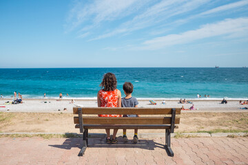 A woman with a child is sitting on a bench on the embankment and looking at the sea