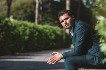 Young thoughtful man sitting with joined hands. Wearing a jacket and trousers. Trendy hair and goatee. Outdoors in a park.