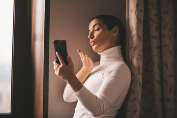 A pretty young smiling Caucasian woman in a white turtleneck sends a kiss and video chat on her mobile phone at home. A woman who has a video call on her smartphone. Video chat in social networks.