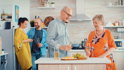Cheerful old elderly adult family hugging laughing dancing drinking a glass of white wine in front young couple. In the kitchen. Grandma and grandpa embracing smiling and daughter taking a photo