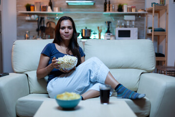 Young lady in pijamas watching Tv in living room sitting on sofa. Bored, home alone late at night...