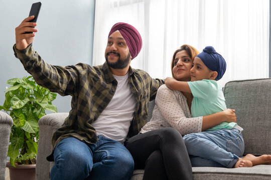 A SIKH SON AND MOTHER HUG EACH OTHER WHILE FATHER CLICKS A SELFIE OF EVERYONE SITTING TOGETHER ON A SOFA	