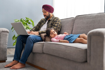 A SIKH MAN WORKING ON LAPTOP WHILE DAUGHTER COMFORTABLY SLEEPING NEAR HIM ON SOFA	