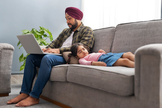 A SIKH MAN WORKING ON LAPTOP WHILE DAUGHTER COMFORTABLY SLEEPING NEAR HIM ON SOFA	 - Powered by Adobe