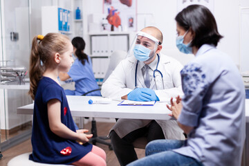 Fototapeta premium Doctor wearing face mask during global pandemic talking with child during consultation. Health pediatrician specialist providing health care services consultations treatment in protective equipment.