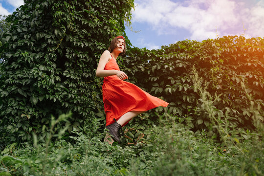 Beautiful Young Rebel Brunet Girl In Red Dress In Green Park Bushes Walking In Black Shoes. Color Contrast Expressive Fashion Style Move. Conceptual Artistic Horizontal Composition Stylish Brutal Teen