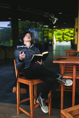 Young black haired man witn piercing reeding a book and smoking tabacco pipe in outdoor cafe