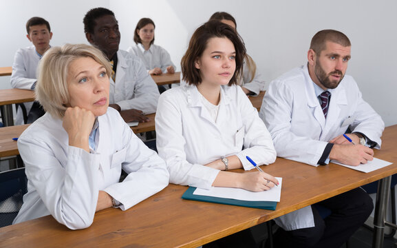 Multinational group of people in white coats attentively listening and making notes while sitting in boardroom