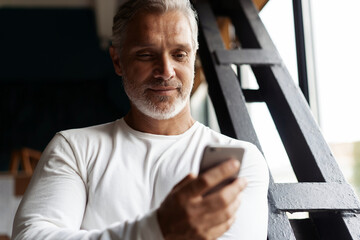 Casual Grey-haired Mature businessman using smartphone standing by the window