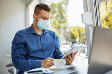 Young businessman working remotely from cafe on a laptop and tablet with a cup of coffee. Leading business during covid-19 pandemic quarantine and staying healthy concept.