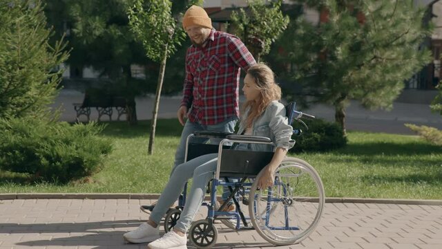 Touching moment of help and support. Young man tie shoelaces to his disabled invalid girlfriend on wheelchair. Couple in love walking outside. Concept of disability assistance. 6k downscale 10 bit