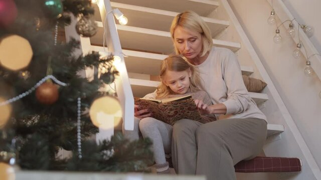 Caucasian female parent sitting on stairs behind Christmas tree and decorative lights, holding kid in arms and reading funny story from book