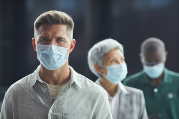 Portrait of young man in protective mask looking at camera while standing in a queue with other people