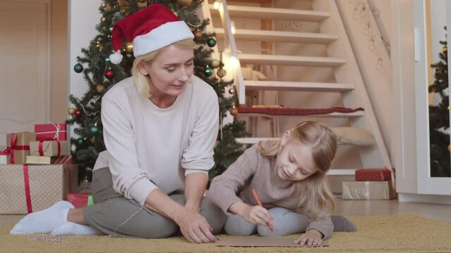 Caucasian blond mother sitting on carpet wearing Santa hat and watching her little daughter draw picture at home in Christmas decorations