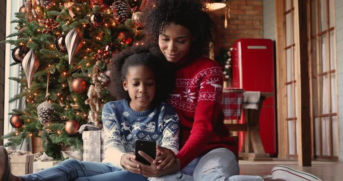 African mother and daughter sitting on floor near glowing decorated Christmas tree using smart phone choose and buy New Year gifts for family on internet. E-commerce retail services remotely concept
