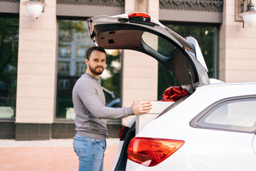 Bearded young delivery man standing near car, large festive white box with beautiful red bow at truck, looking at camera. Courier preparing to take parcel out of trunk of car and deliver to customer.