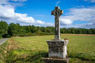 Old stone grey cross standing against background scene of green forest and field at the road crossing