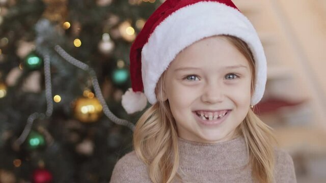 Portrait of cheerful blond blue-eyed Caucasian little girl wearing red Santa hat looking and smiling on camera, Christmas decorations in background