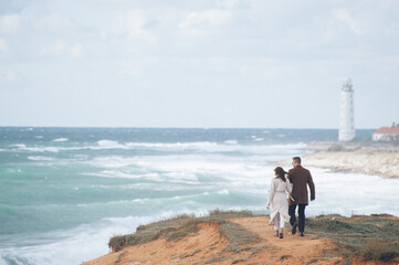 two young people man and woman in coat walking along sea shore with lighthouse during windy storm weather in winter