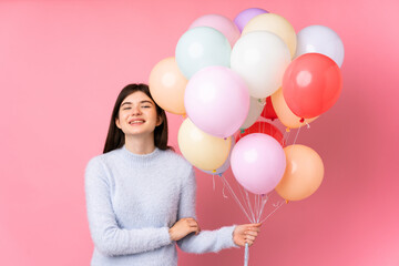 Young Ukrainian teenager girl holding lots of balloons over isolated pink background laughing