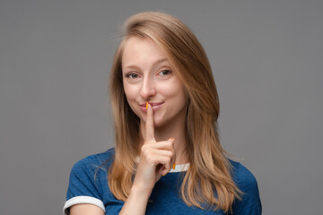 Pretty young blonde woman, dressed in blue t shirt, keeps finger on lips, making hush gesture on gray background. Shh, silence concept