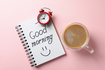 Notebook with text Good morning, clock and cup of coffee on pink background