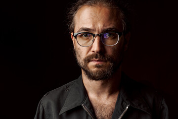 Dark studio portrait of a middle-aged man with beard in glasses