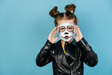 Fototapeta premium Halloween holiday or Day of Death concept. Angry little girl with serious expression, keeps hand near face, wears creative makeup, dressed in black leather jacket, isolated over blue background