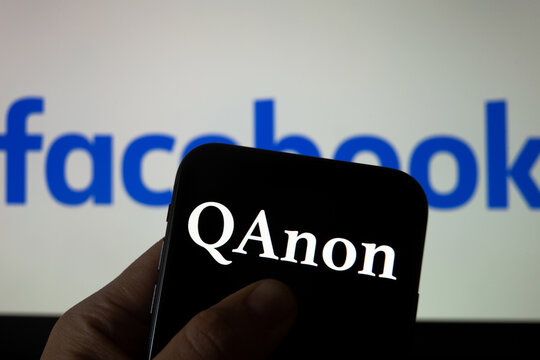 QAnon vs FACEBOOK. QAnon organization logo seen on the smartphone which is placed on Facebook logos. Concept for ban of QAnon on social media. 