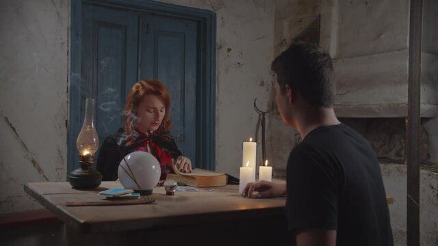 Concentrated attractive female tarot reader fortune telling to worried man, interpreting tarot cards with ancient spellbook and telling bad news while predicting the future in rustic house at dusk.