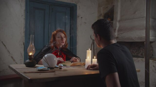 Mysterious young red haired soothsayer predicting destiny with ancient magic book, reading tarot cards and forecasting future to curious young man while fortune telling in rural farmhouse at dusk.