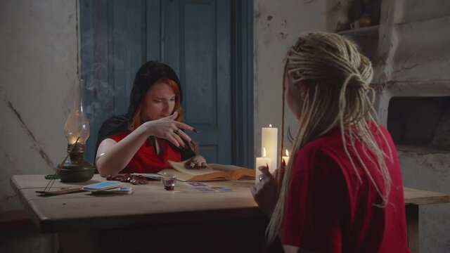 Pretty professional fortune teller with magic book tarot card reading, forecasting the future, telling to worried young woman bad prediction while practicing divination in rustic house in the evening.