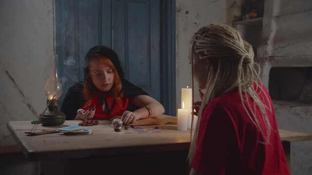Concentrated charming female fortuneteller in black cape casting magic runes on rustic table, predicting and interpreting the future with spellbook to worried woman during divination in rural house.