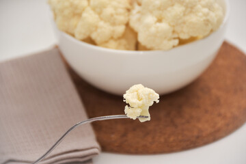 A piece of cauliflower on a fork with cauliflower in a white bowl in the background.