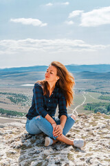 Beautiful young brunette woman in ripped jeans sitting on a cliff overlooking the rustic landscape, White rock in Crimea