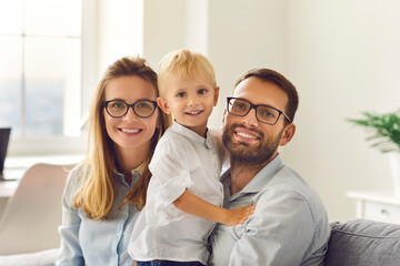 Happy young smiling family sitting, holding little son on knees and looking at camera