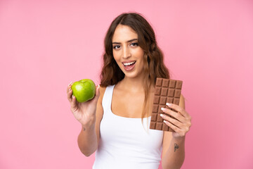 Young woman over isolated pink background taking a chocolate tablet in one hand and an apple in the...