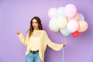 Young woman catching many balloons over isolated on purple background surprised and pointing finger to the side